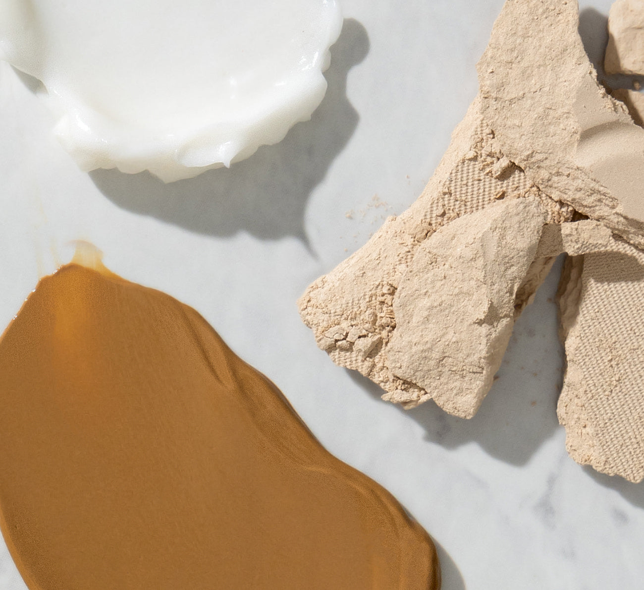 Smear of tan liquid foundation, dollop of white cream, and crumbled beige pressed powder on marble surface.