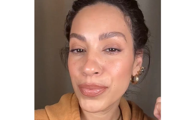 Close-up of woman with glossy nude lipstick, subtle blush, defined brows, and gold hoop earrings.