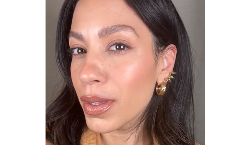Close-up of woman's face showing glossy nude lipstick, warm bronzer, and a textured gold hoop ear cuff.