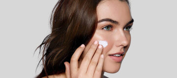 Young woman smiling as she applies white facial moisturizer to her right cheek