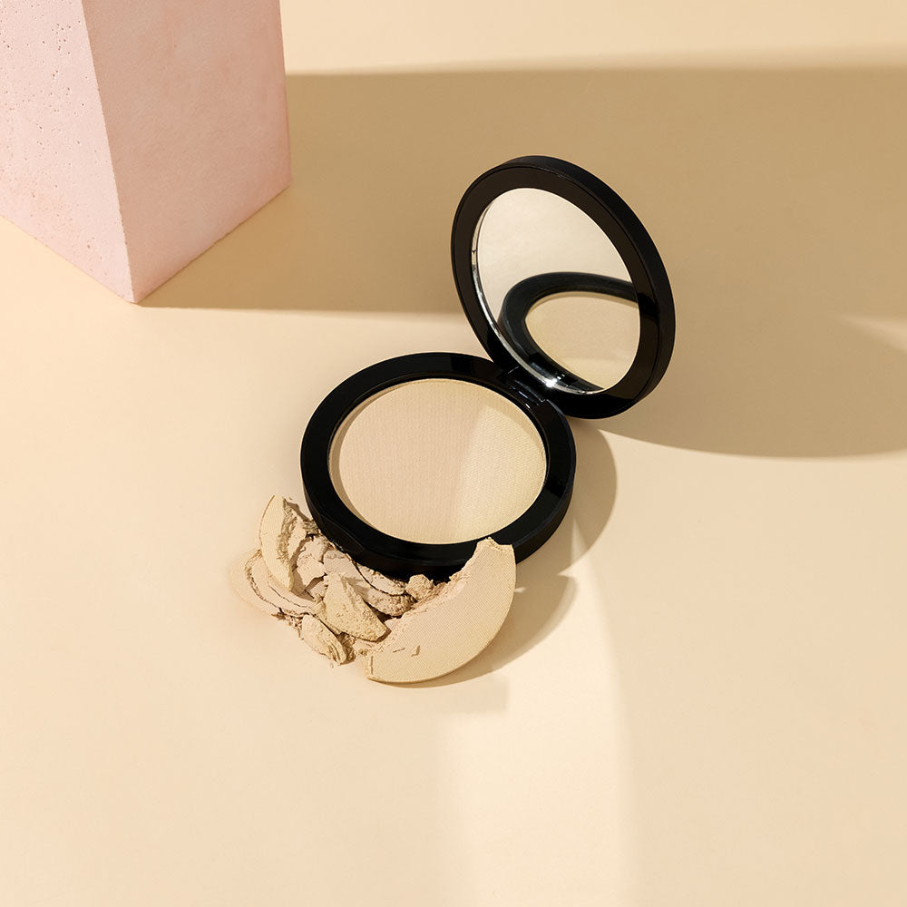 Open compact powder with mirror and crushed pale-beige powder on a beige surface