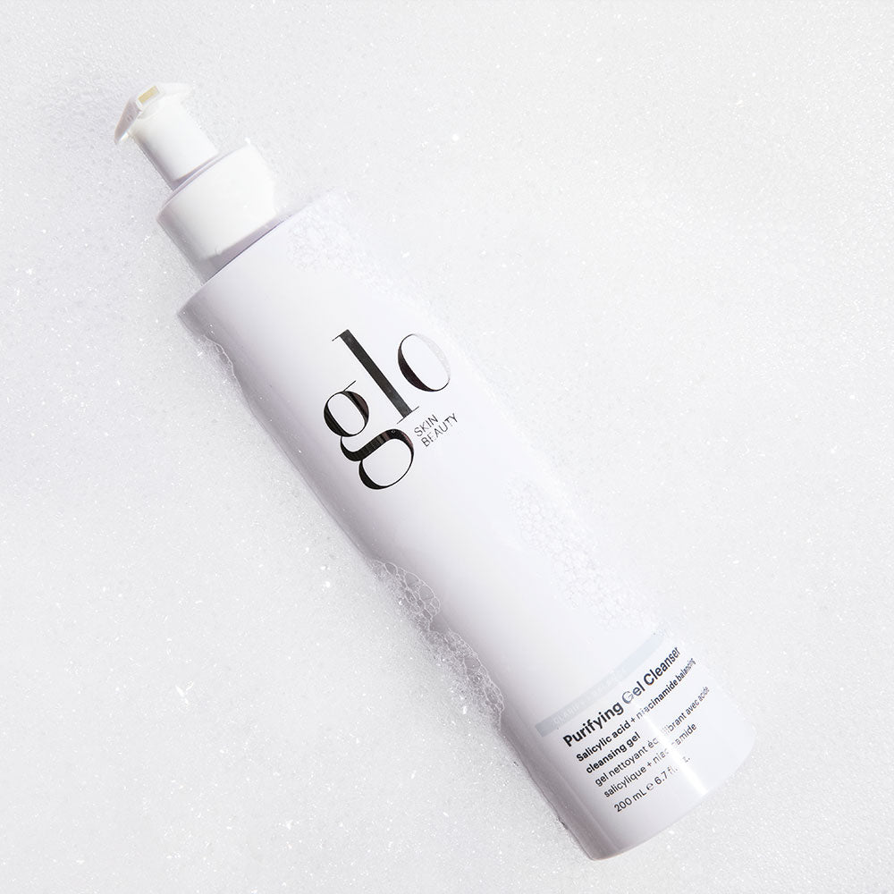 glo SKIN BEAUTY Purifying Gel Cleanser bottle with pump resting in white foamy bubbles