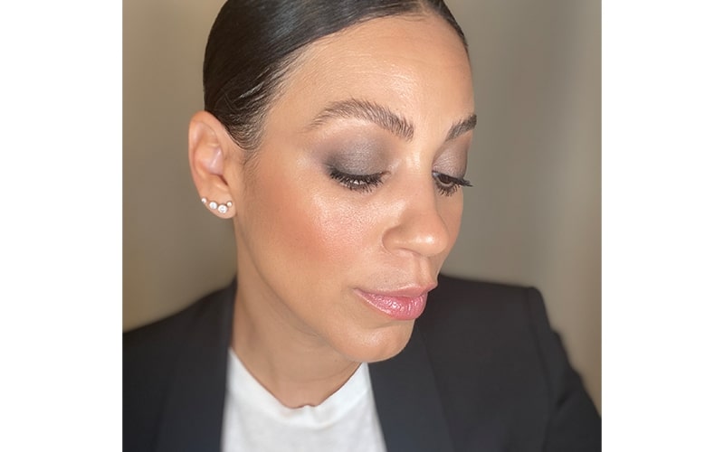 Woman side profile with dark smokey eye makeup, glowing skin, stud earrings, wearing a white top and black blazer.