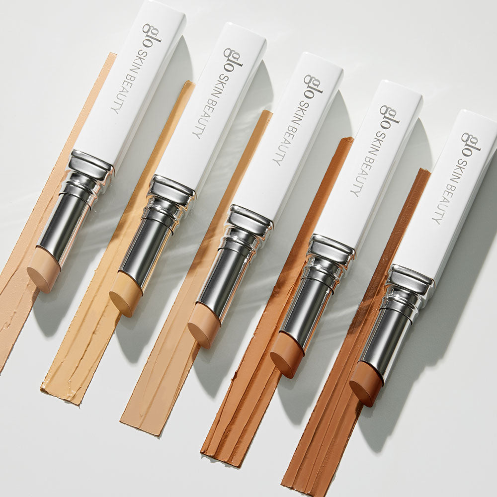 Five glo SKIN BEAUTY stick concealers in graduated light-to-deep shades, each with a swatch streak beneath.