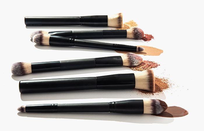 Five black-handled makeup brushes arranged diagonally with foundation and powder swatches on a white background