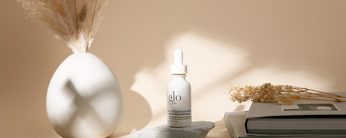 White dropper bottle labeled 'glo' and 'Beta-Clarity BHA Drops' on a stone, beside a white vase and dried flowers on a beige background