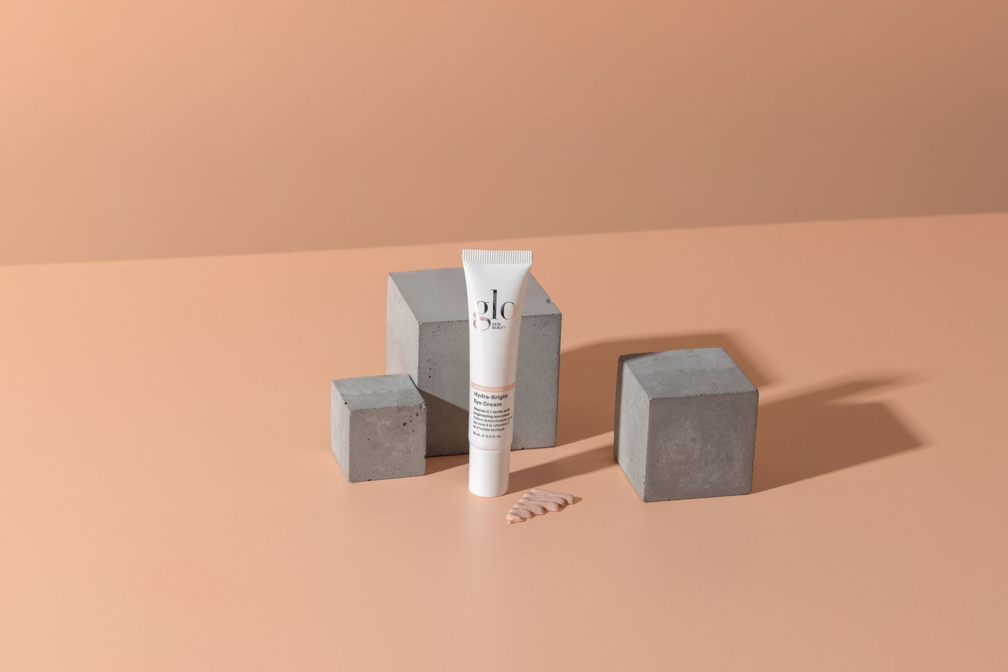 glo Hydra-Bright Eye Cream tube on peach background with concrete blocks and a dab of cream