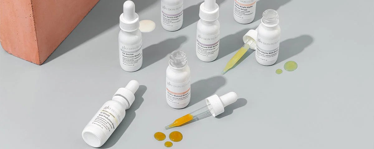 glo SKIN BEAUTY dropper bottles on a gray surface with scattered yellow and green serum droplets