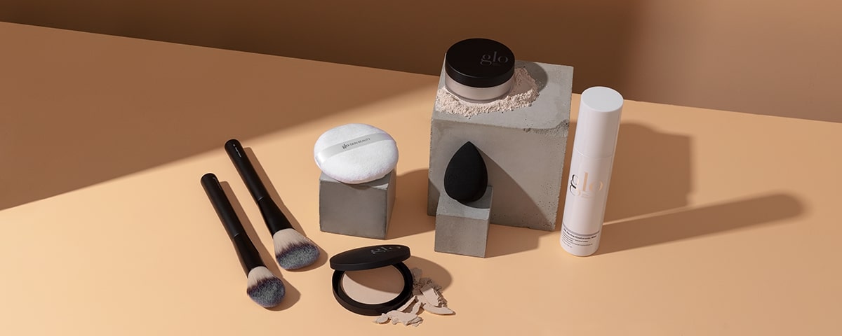 Pressed compact and loose powder jar (glo) with brushes, powder puff, and blending sponge on a beige surface