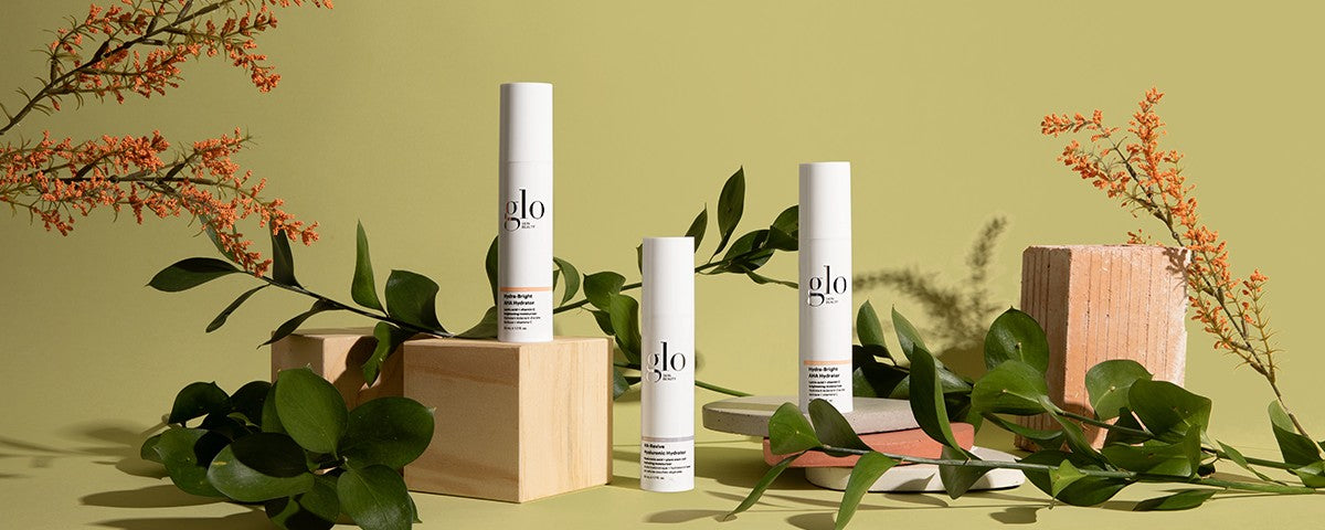 Three white Glo skincare bottles on wood blocks with green leaves and orange sprigs against a pale green background.