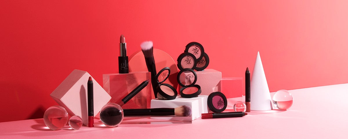 Assorted glo blush compacts, lipstick, brushes, and pencils arranged on a pink backdrop with geometric display props