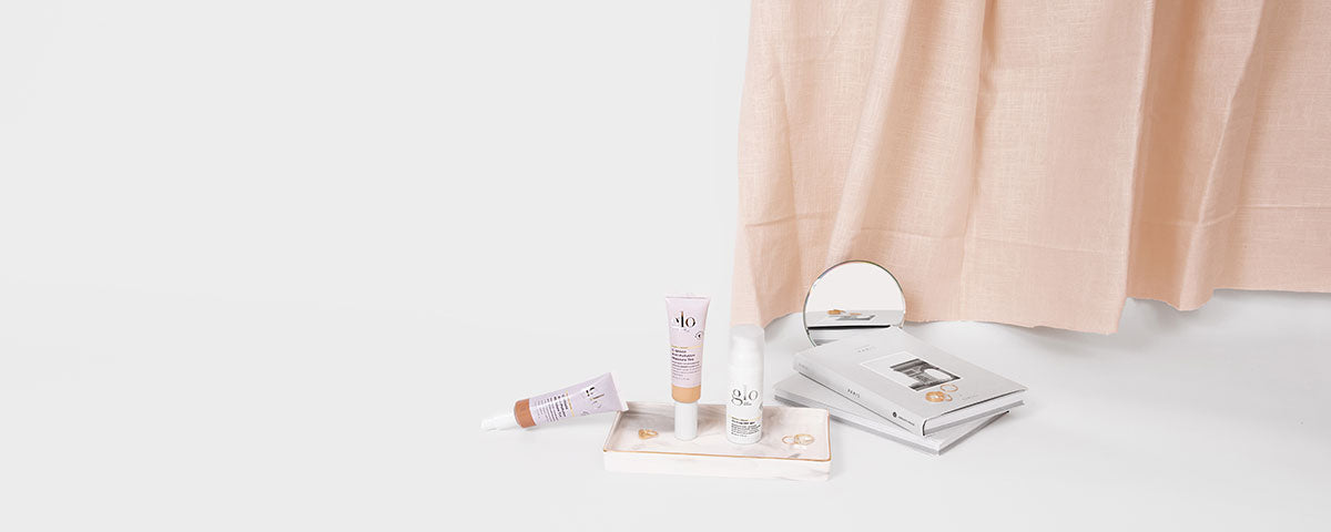 Three glo skincare tubes and a pump bottle on a small tray beside stacked booklets and a round mirror under a pink curtain
