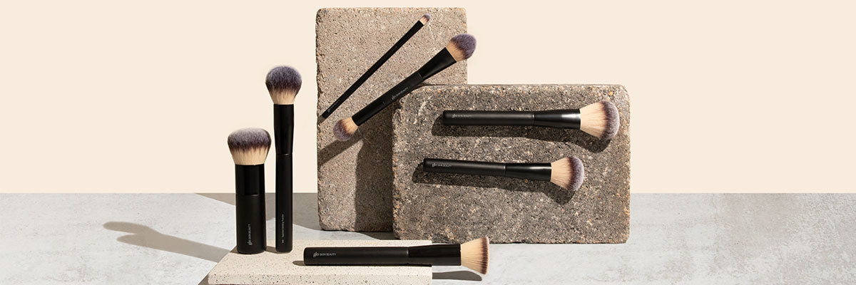 Six black-handled makeup brushes with rounded and flat bristles arranged on textured stone blocks against a beige background.