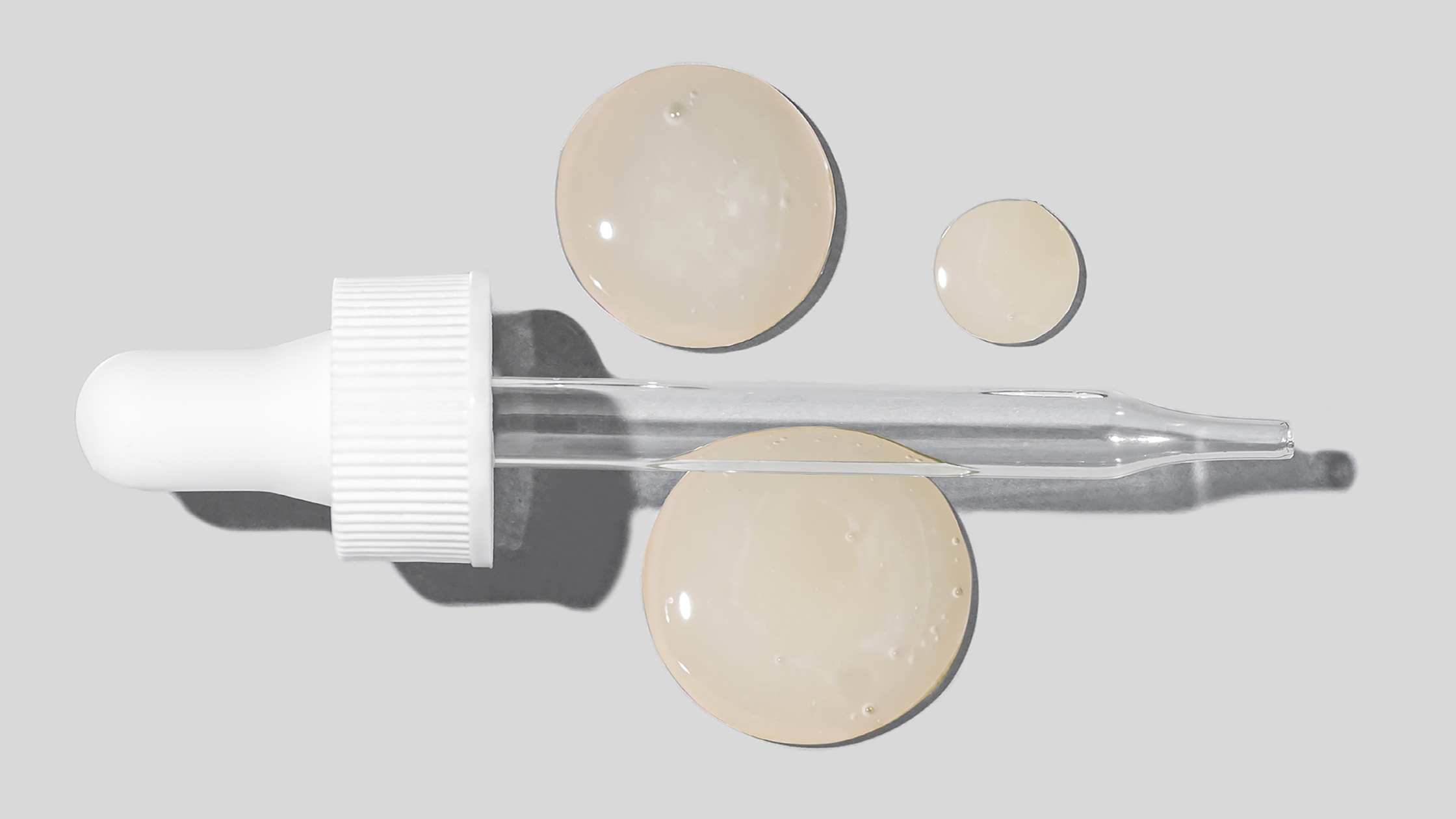 Glass dropper with white rubber top and three beige serum droplets on light gray background
