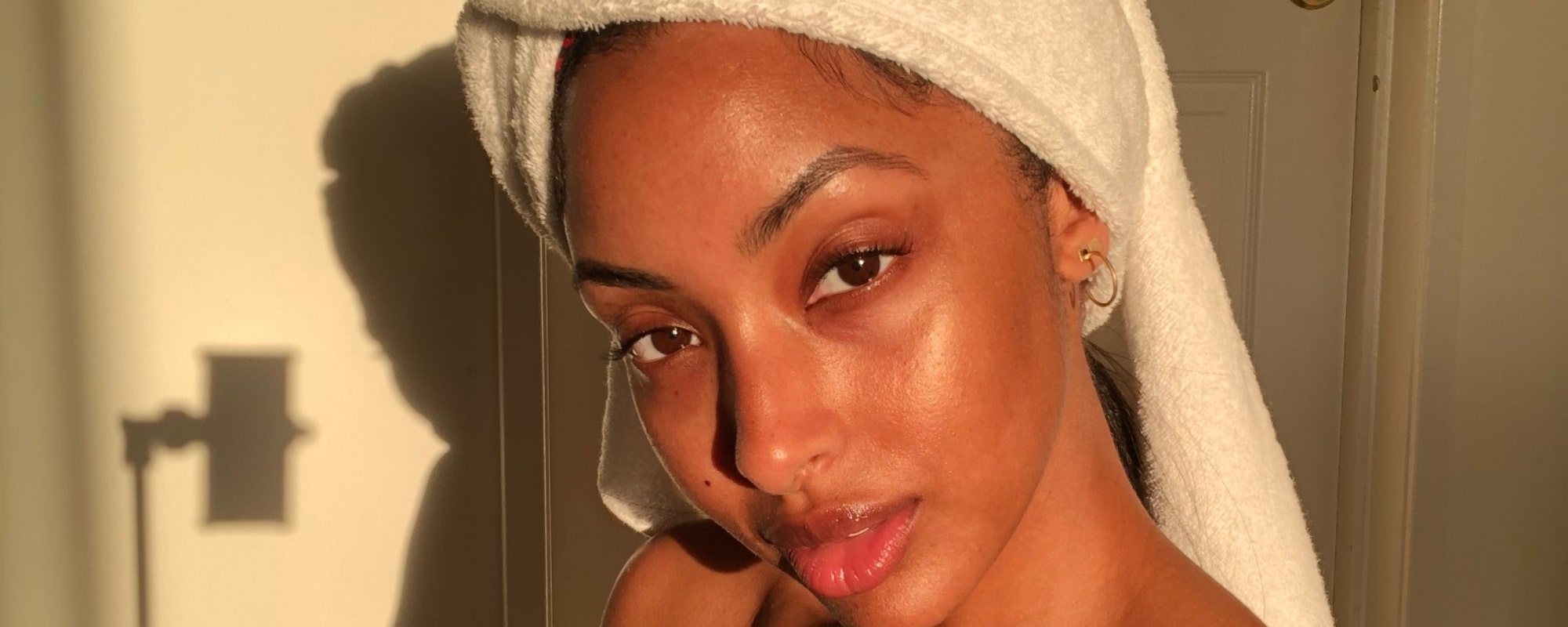 Close-up portrait of a woman with towel-wrapped hair and dewy skin, lit by warm side sunlight.