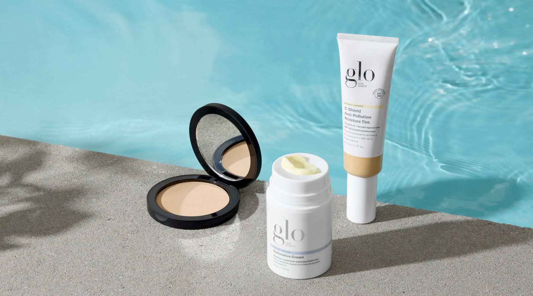 Front view of three Glo Skin Beauty products: open pressed powder compact, restorative cream jar with cream on top, and C‑Shield Anti‑Pollution Moisture Tint tube by a pool