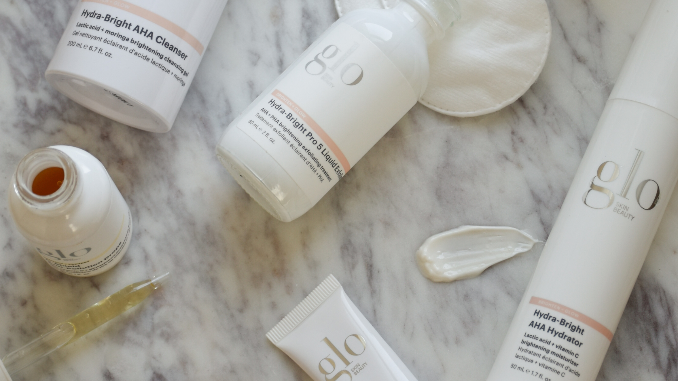 glo Skin Beauty Hydra-Bright AHA Cleanser; Pro 5 Liquid Exfoliant; AHA Hydrator — flat lay on marble with cream smear and cotton pad