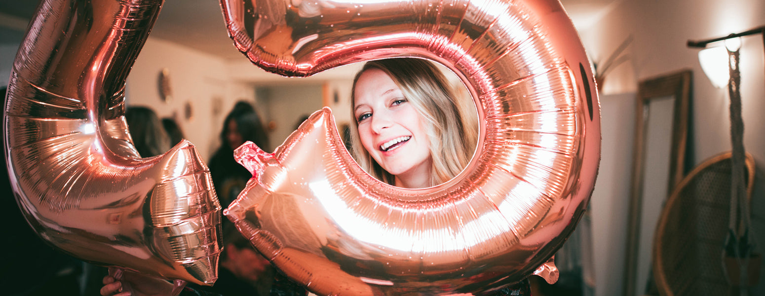 Smiling woman peeks through a rose-gold foil number 5 balloon at a small indoor party.
