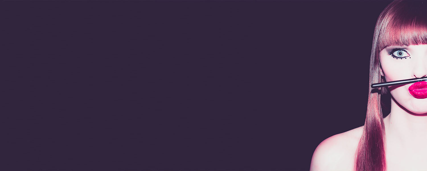 Woman's right face with straight bangs, bright fuchsia lipstick, and a makeup brush held horizontally across her mouth