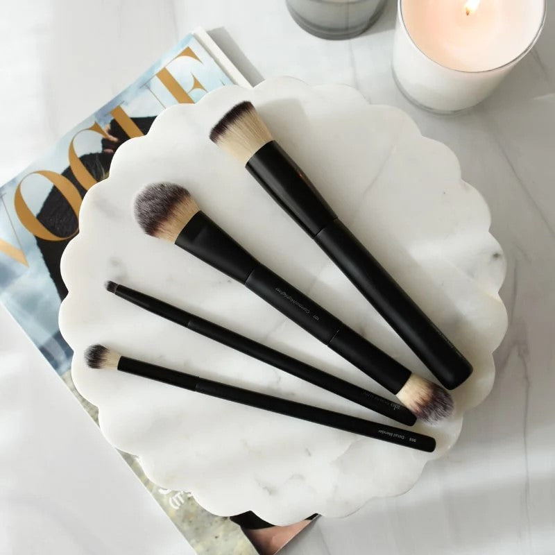 Five black-handled makeup brushes arranged on a scalloped white marble tray over a VOGUE magazine next to a lit candle