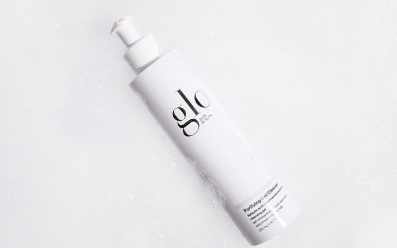 White pump bottle labeled 'glo skin beauty' and 'Purifying Gel Cleanser' resting on foamy bubbles