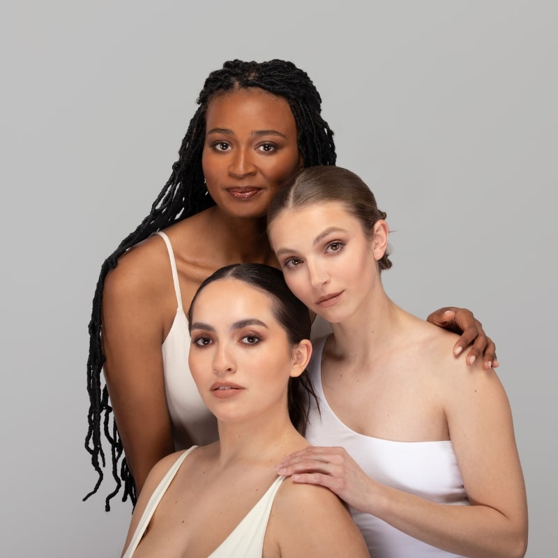 Three women with different skin tones wearing neutral tops, close together showing contoured makeup on faces.