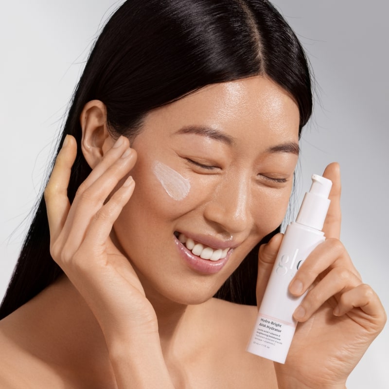 Smiling woman rubbing cream on cheek while holding bottle labeled glo Hydra-Bright AHA Hydrator.