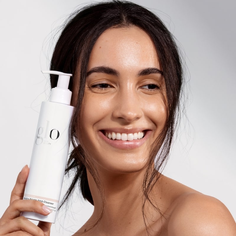 Smiling woman holding a white pump bottle labeled glo cleanser near her cheek.