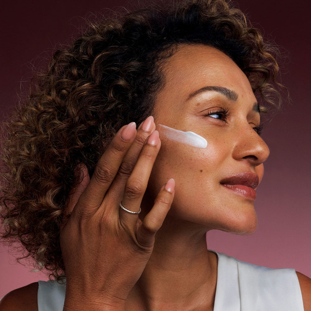 Woman applying Bio-Renew EGF Cream to her cheek against a fig-colored gradient background