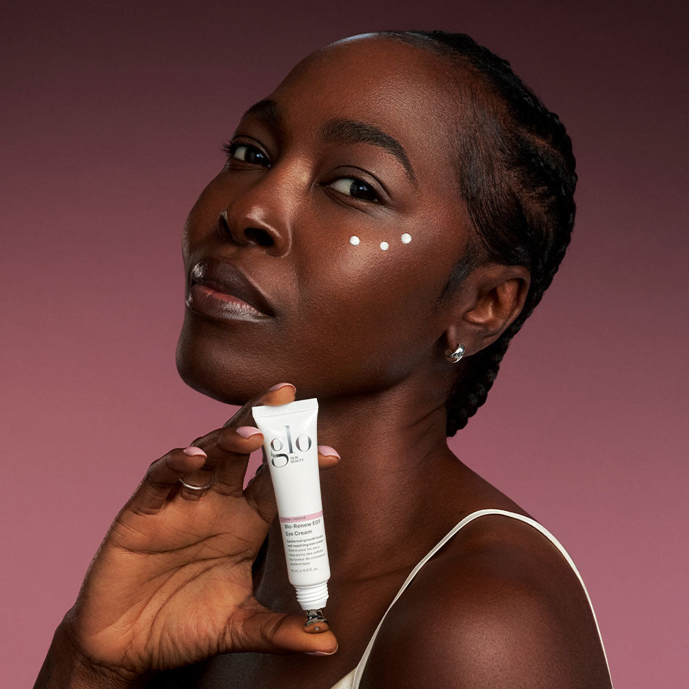 Handheld Bio-Renew EGF Eye Cream tube with metal roller applicator and dots of cream on model’s cheek, pink background