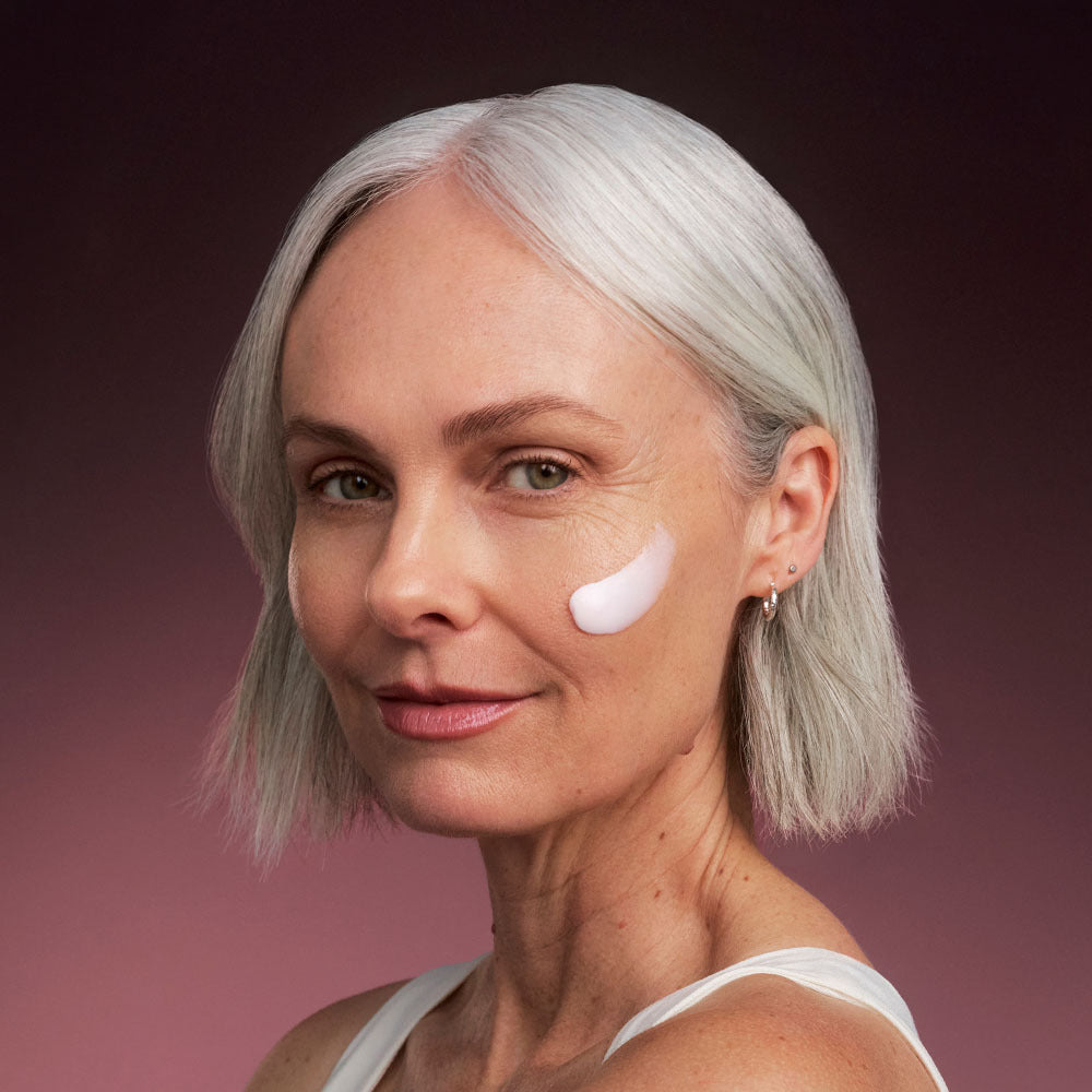 Older woman with a swipe of pale pink repair cream on her cheek, looking at camera against a dusky pink background