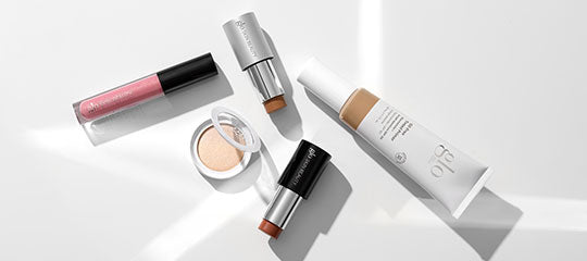 Assorted makeup products arranged on a white surface: foundation tube, cream compact, two lipsticks, and a pink lip gloss