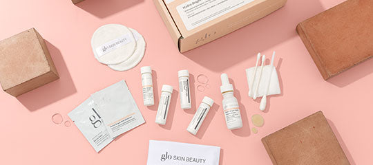 Assorted glo SKIN BEAUTY sample skincare products and applicators on pink background: vials, dropper bottle, sachets, cotton pads, boxes.