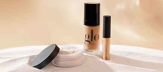 Loose powder jar, liquid foundation bottle labeled glo, and concealer tube arranged on textured beige fabric.