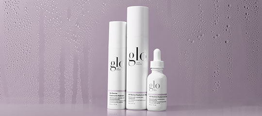 Three white 'glo' skincare product bottles on a purple background with water droplets