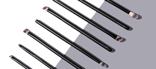 Eight black-handled eye makeup brushes with angled bristles arranged diagonally on a split white and gray background.