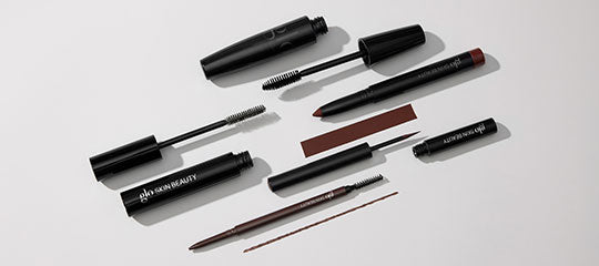 Assorted black eyebrow and mascara tools, including a tube labeled 'glo SKIN BEAUTY', arranged on a light background.