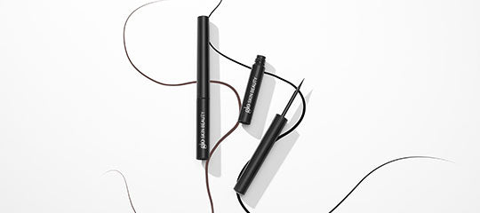 Three black mascara tubes with a detached wand and curved brown and black eyeliner strokes on white background