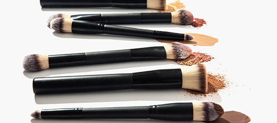 Five black-handled makeup brushes with foundation swatches and loose powder on a white background