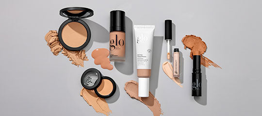 Assorted glo makeup: liquid foundation, concealer, stick concealer and pressed powders with swatches on light gray background