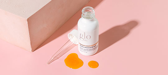 White bottle labeled 'glo' with dropper and three spilled orange serum drops on pink surface beside a white block.