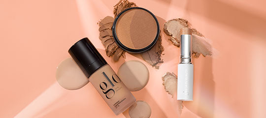 glo foundation bottle, bronzer compact with powder swatches, and a white lipstick tube on peach background.