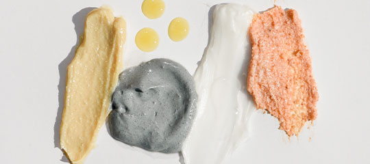 Four cosmetic product swatches in beige, gray, white, and peach with three yellow droplets on a white background