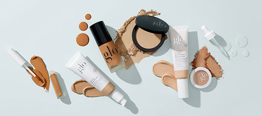 Assorted glo cosmetics: foundation bottles, compact, tubes and cream jar with swatches on pale blue background