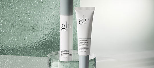 Two Glo Skin Beauty skincare tubes labeled Microbiome Restoring pH Moisturizer and Dual Clarifying Cleanser on a glass surface.