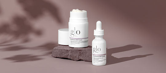 Two white glo skincare containers, a jar and a dropper bottle, on a mauve surface with soft leaf shadows