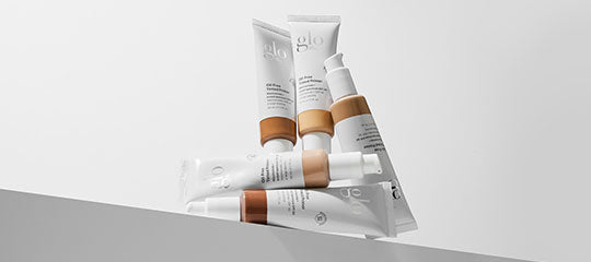 Five glo skincare tubes in white packaging with tan caps arranged on a gray ledge, showing 'glo' branding.