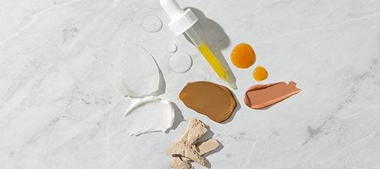 Skincare product textures and makeup color swatches: white cream, beige powder, brown and peach balms, amber serum drops and a glass dropper on marble