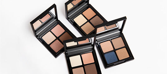 Four open quad eyeshadow palettes showing neutral and blue shades in black compact cases with mirrors on white background.