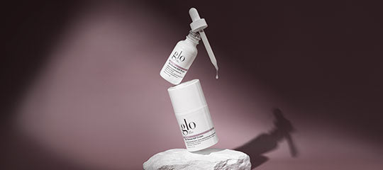 Two Glo Skin Beauty skincare products, one with a dropper, displayed on a stone pedestal with a purple background.
